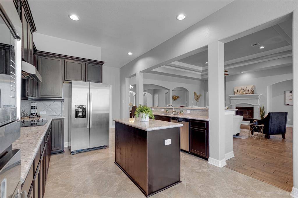 This is a spacious, open-concept kitchen with modern stainless steel appliances, dark wood cabinetry, and granite countertops. It flows seamlessly into a cozy living area with a classic fireplace, highlighted by elegant archways and tray ceilings.