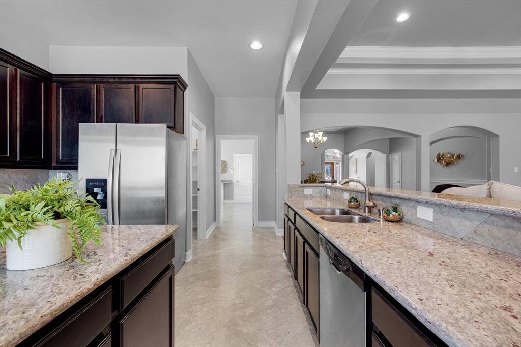 This modern kitchen features dark wood cabinetry, stainless steel appliances, granite countertops, and tile flooring. The open layout connects to a living area with arched wall openings, offering a spacious and inviting atmosphere.