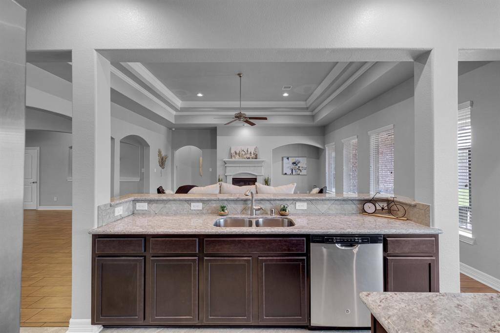 This image showcases a modern, open-plan kitchen with dark wood cabinetry, granite countertops, and stainless steel appliances. The space is well-lit, featuring a tray ceiling with recessed lighting and natural light from multiple windows with blinds. The kitchen overlooks a comfortable living area, providing a seamless flow for entertaining and daily living.