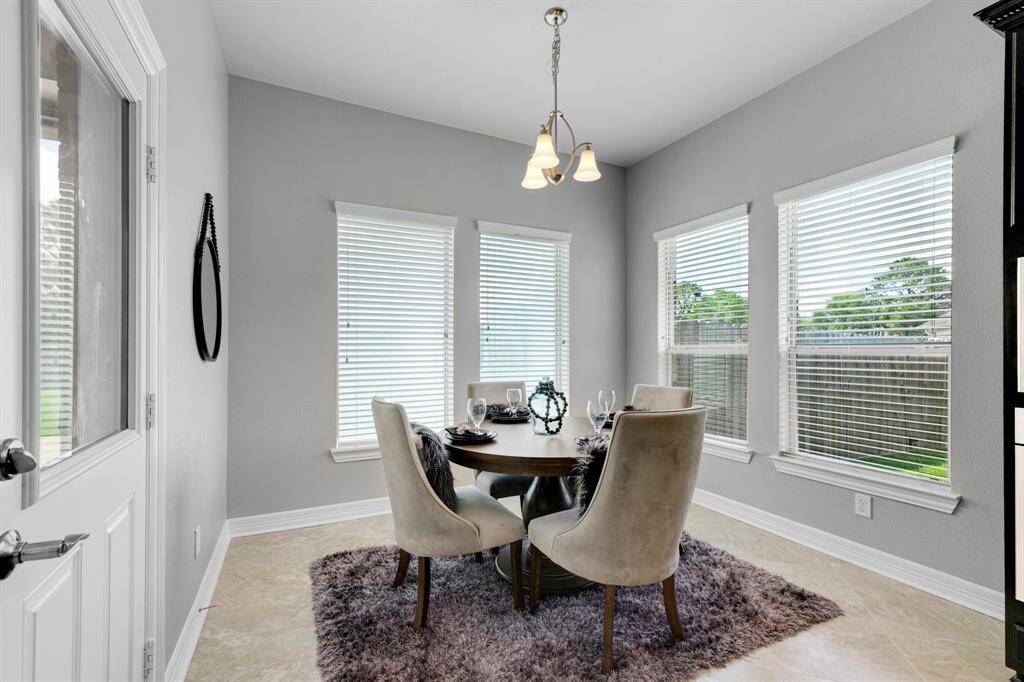 This is a modern dining area featuring a sleek round table with comfortable upholstered chairs, set on a plush area rug. Natural light streams in through two large windows with blinds, and the space is accented by a simple yet elegant light fixture. The room is painted in a neutral gray tone, which complements the light tile flooring.