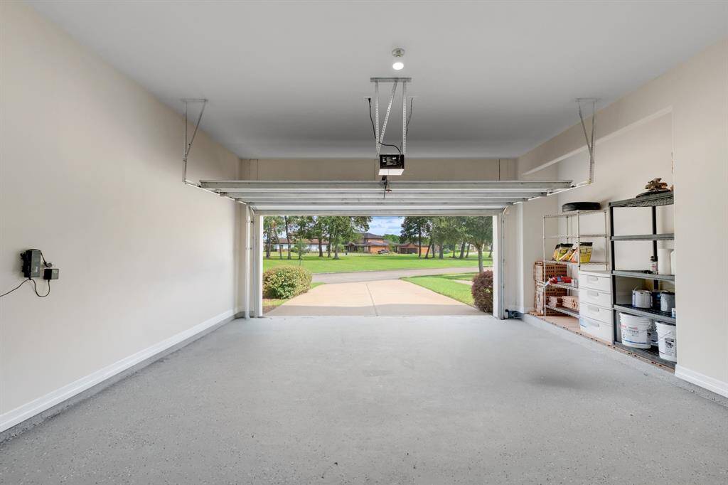 You're looking at a spacious, clean two-car garage with an automatic door opener and built-in shelving for storage. It opens up to a well-kept driveway and offers a view of a grassy area with trees.