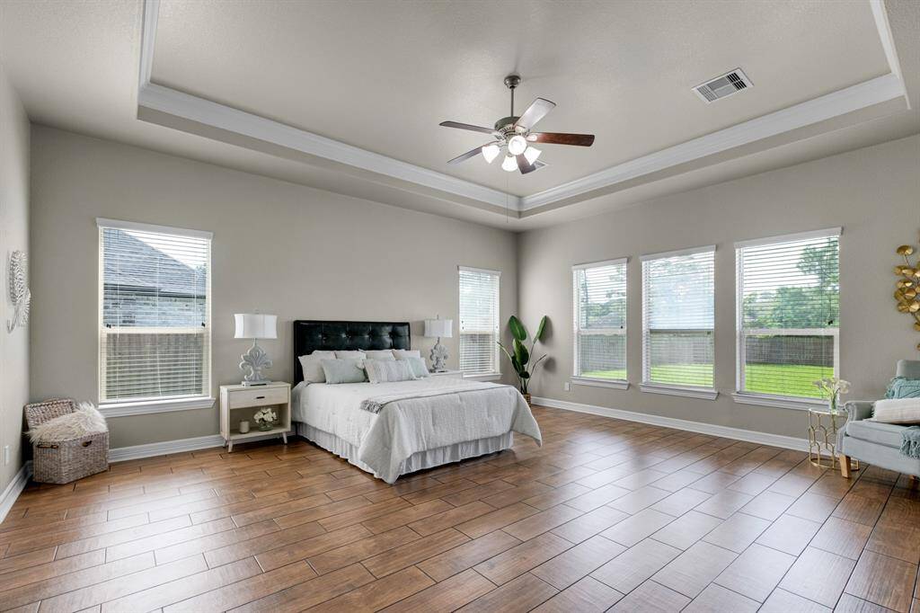 This is a spacious and well-lit master bedroom featuring a tray ceiling with a ceiling fan, large windows with a view of the yard, and elegant wood flooring. The room is neutrally decorated, offering a serene and versatile space.