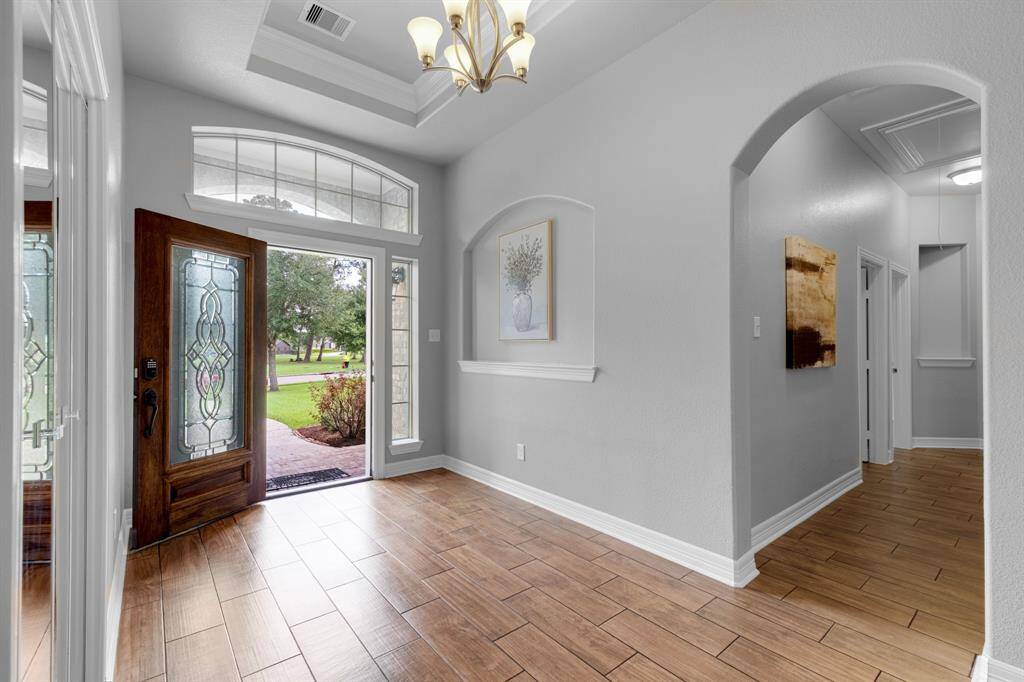 This is a bright and welcoming entryway with high ceilings, a decorative glass front door, transom and sidelight windows, and wood-look tile flooring. The space features arched doorways and neutral colors, offering a modern and elegant feel.