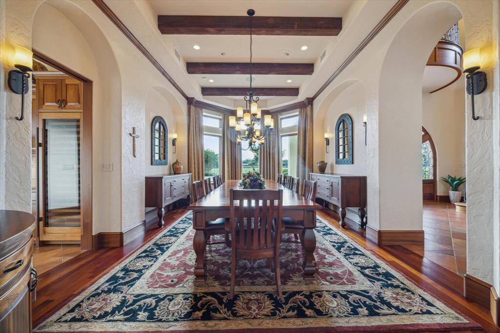 The intimate formal dining space comfortably accommodates a table for 10-12 dinner guests. A tray ceiling with wood beams accentuates the Mediterranean architecture. Wrought iron chandeliers and sconces create a pleasing ambiance. The bay of windows with transoms provides a lovely view of the courtyard and like all windows in the home, feature Lutron electronically powered shades. Sideboards nestle into the dedicated arched niches and a built-in corner cabinet provides additional storage.