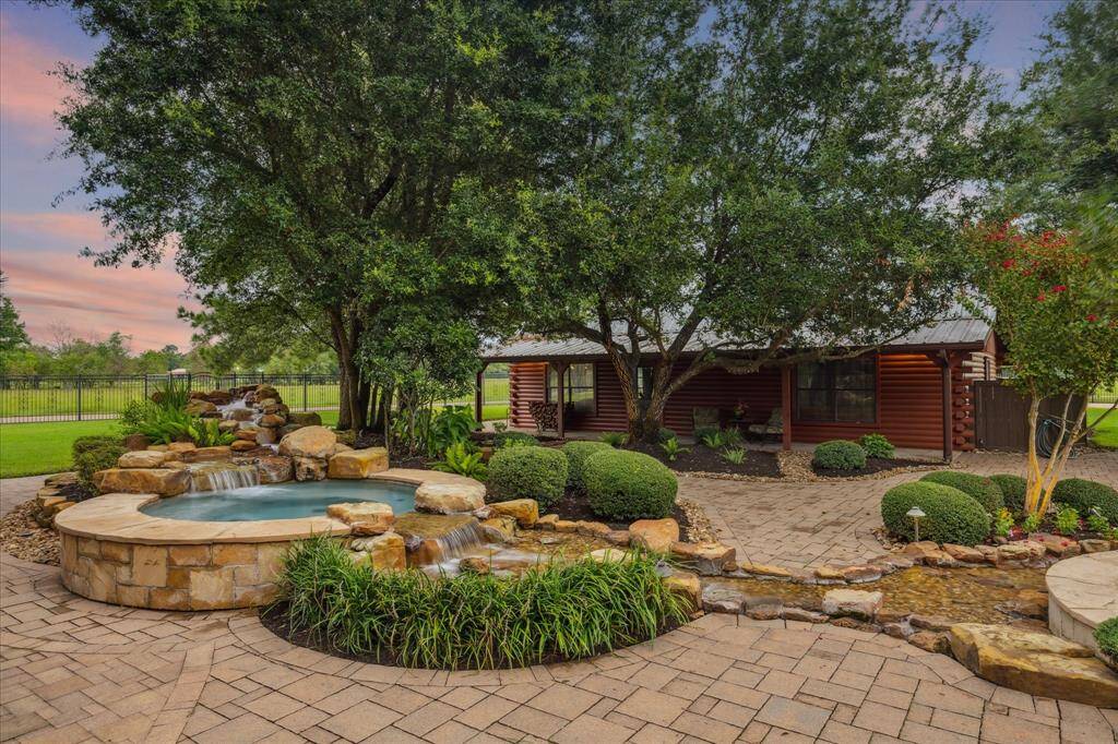 The hot tub is intentionally separated from the main pool, connected by a waterfall and rock stream and located just steps away from the primary bedroom suite. In the background, the log cabin guest house is visible, nestled beneath the shade of the mature trees.