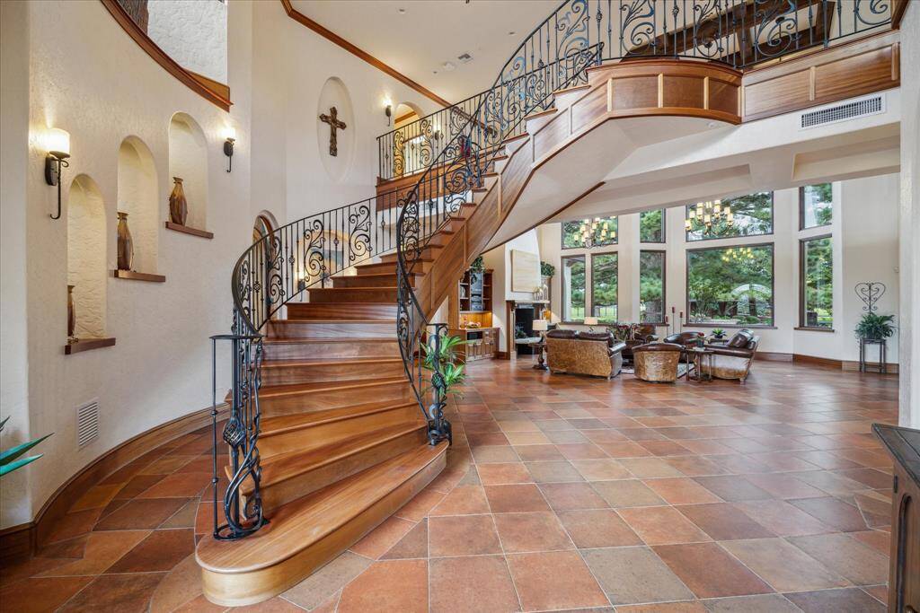Inside, a soaring 26’ ceiling and the sweeping, single-piece solid wood staircase with decorative, custom wrought iron railing leads the eye to the upper levels. The custom railing is one single wrought iron piece. Aesthetically, the crisp white painted walls are a minimalist backdrop complemented by the lush layering of materials throughout the home as seen here with the Saltillo tile floor, richly stained mahogany wood and the iron railing.