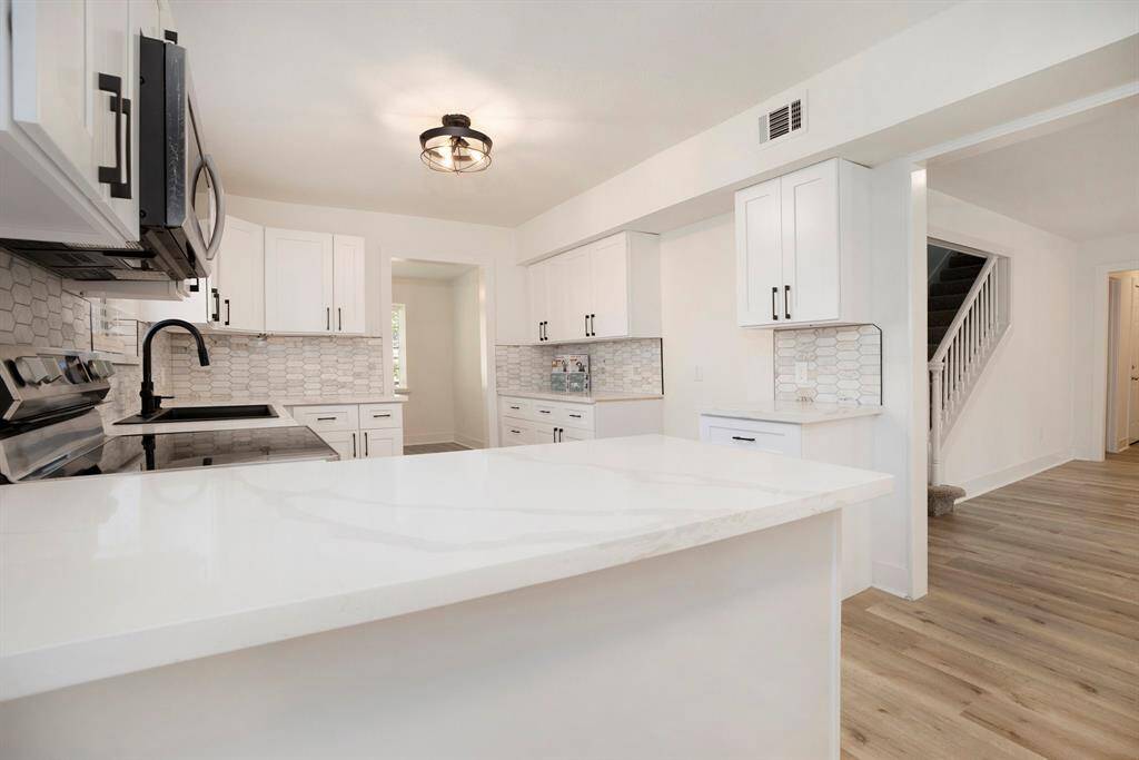 Modern kitchen with white cabinetry, stainless steel appliances, sleek subway tile backsplash, quartz countertops, and light floors. Bright, open space with a view of the staircase.
