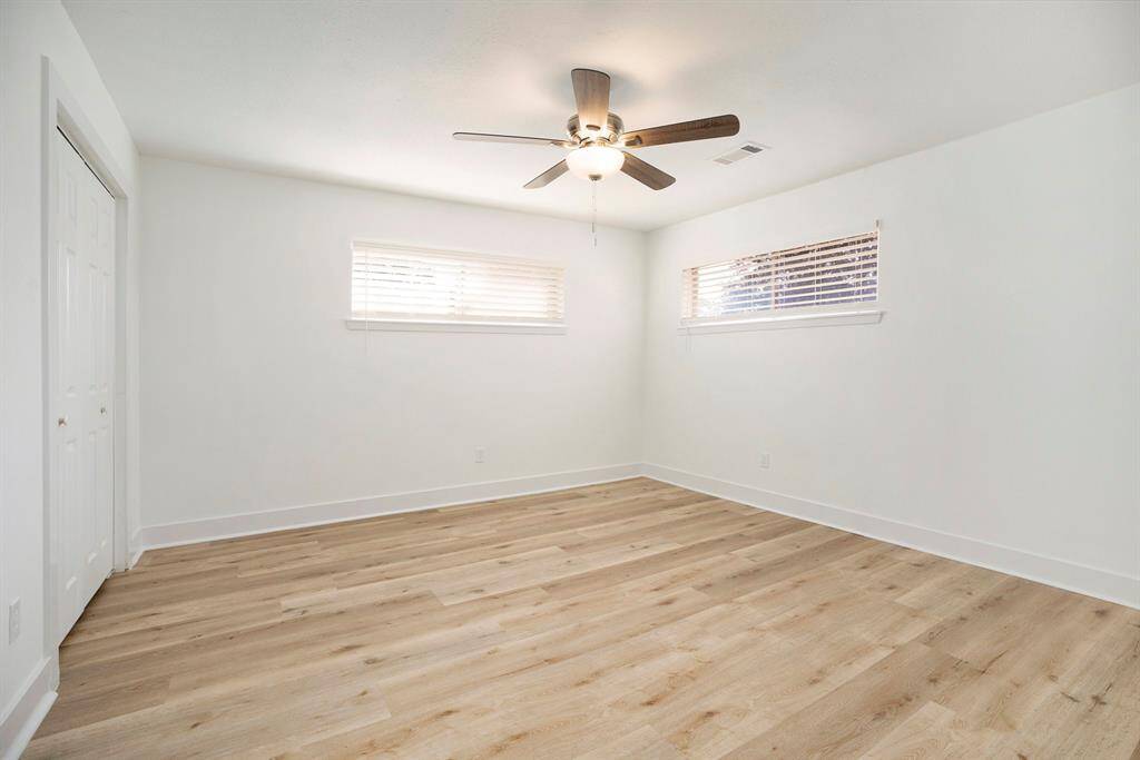 This is a bright primary bedroom featuring light flooring, fresh white walls, two windows with horizontal blinds, and a ceiling fan with lights.