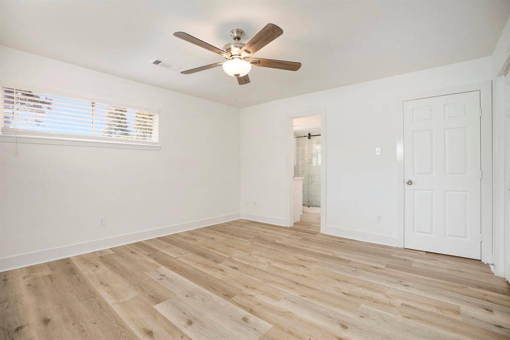 This is a bright primary bedroom room with modern light flooring, fresh white walls, a ceiling fan with lighting, and a large window with horizontal blinds. There's a doorway leading to  an en suite bathroom.