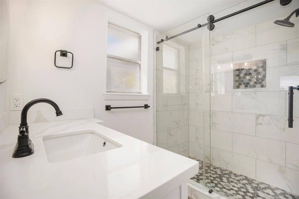This is a modern bathroom with a brand new walk-in shower featuring a glass door and beautiful tiles. There is a single vanity with a white countertop, as well as a frosted window for natural light and privacy.