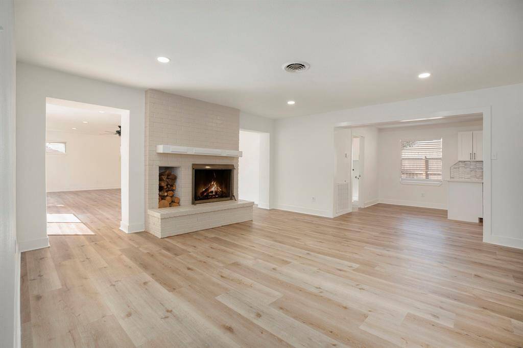 This is a spacious, well-lit room with modern light hardwood floors, featuring a large white brick fireplace with a wood storage nook. It includes multiple openings to adjacent rooms, offering a fluid layout, and a neutral color palette ideal for personalization.