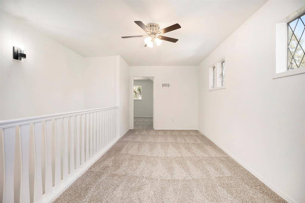 You're looking at a spacious, well-lit upstairs area featuring neutral carpeted flooring, fresh white walls, a ceiling fan for airflow, and charming windows allowing for natural light. The half-wall with a white railing adds an open, yet secure feel, perfect for an upstairs landing or loft area.