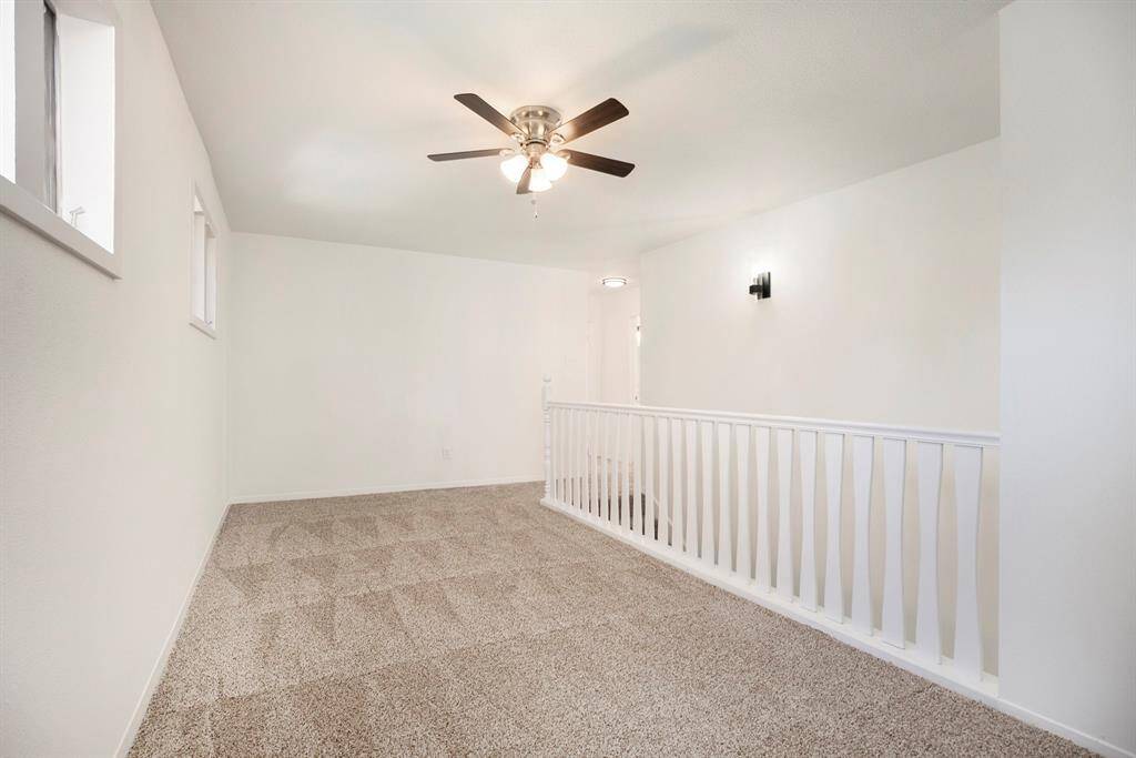 This is a bright, open loft space featuring neutral walls, carpeted flooring, and a ceiling fan. It includes a half-wall railing for safety and overlooks downstairs.