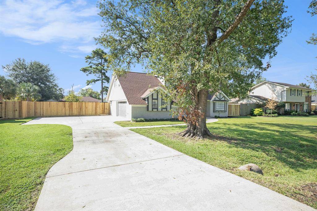 This is a charming home featuring a pitched roof and an attached two car garage, with a wide driveway and a winding path leading to the front door. The property boasts a spacious, well-maintained front lawn with a mature tree, and is fenced for privacy.