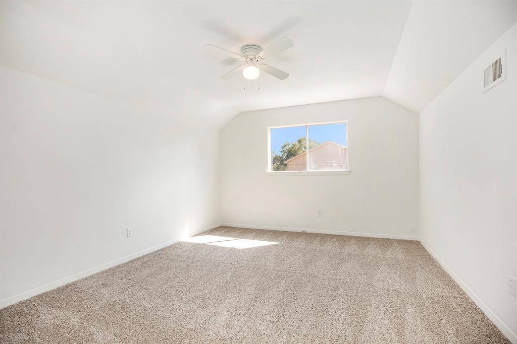 This is a bright secondary upstairs bedroom with fresh neutral paint, plush carpeting, and a ceiling fan. It features a large window providing natural light and a view of the exterior.