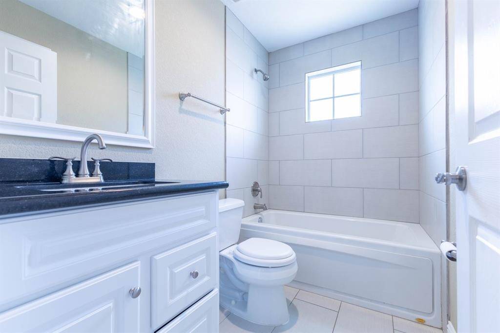 This is a bright, clean bathroom featuring a white vanity with a black countertop, a large mirror, a toilet, and a full tub with a shower. Natural light streams in from a small, frosted window.