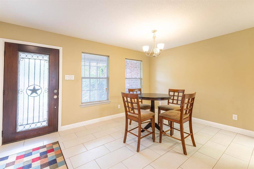 This is a well-lit dining area featuring a decorative front door with glass panels, large windows, a chandelier, complemented by neutral walls and tiled flooring.