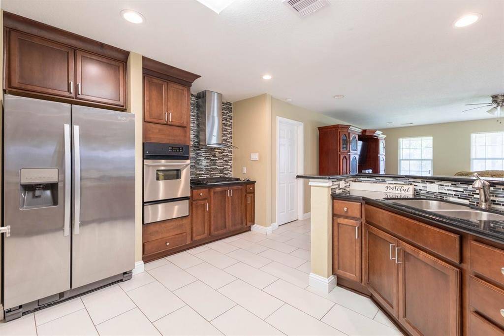 This is a spacious and modern kitchen featuring a built-in oven, rich wooden cabinetry, a stylish backsplash, and a large island with a sink. The tiled floor and neutral color palette offer a clean and inviting ambiance.
