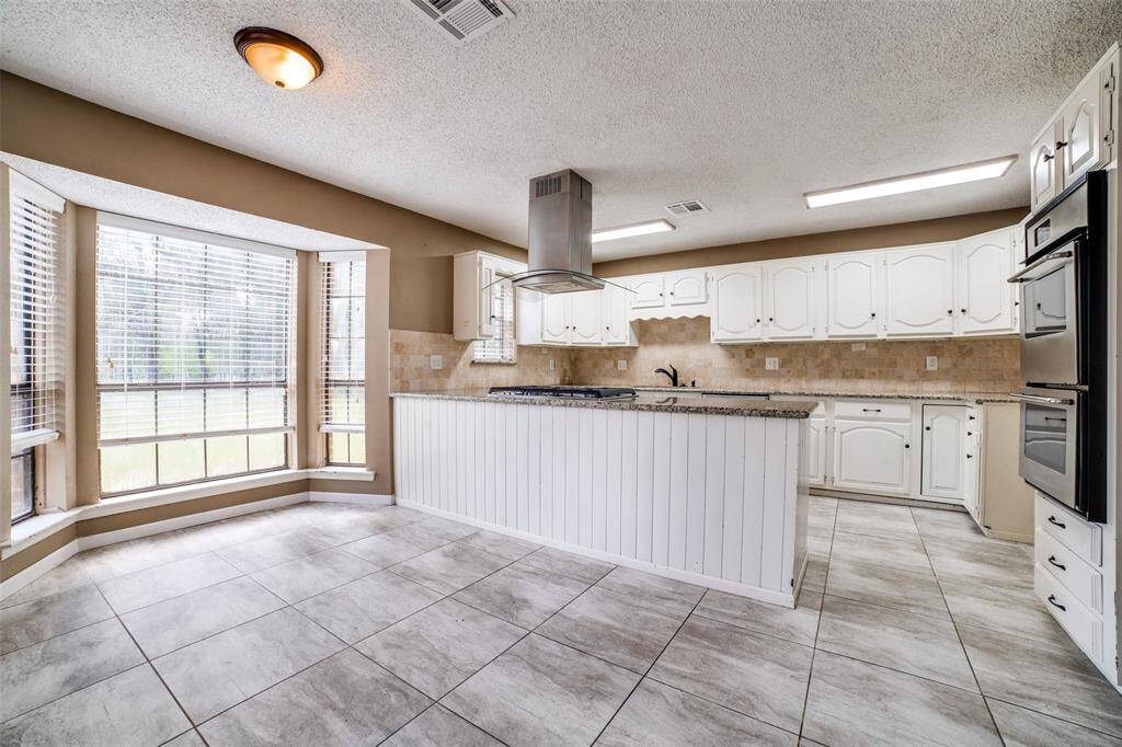 This is a spacious, well-lit kitchen featuring white cabinetry, modern appliances, a tiled backsplash, and large windows with blinds. The room has ample counter space, a central island, and grey tile flooring.