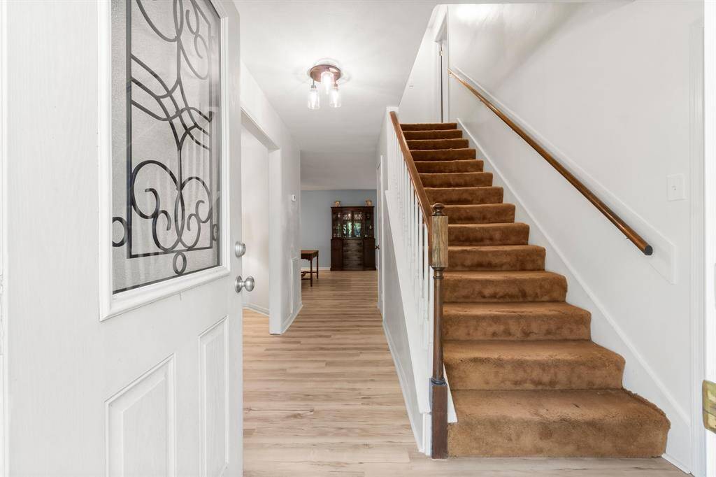 This photo showcases a bright entryway leading to a stairway with plush, tan carpeting. The area features light hardwood floors, a decorative glass-paneled door, and white walls, creating a clean and welcoming atmosphere.
