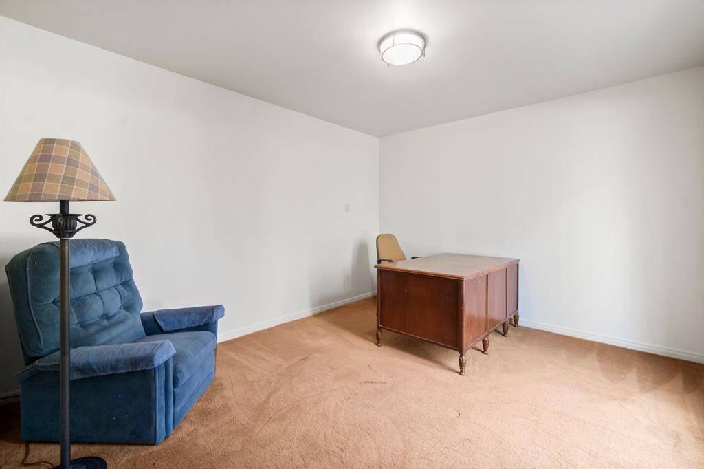This is a modestly sized, unfurnished room with beige carpeting, white walls, and a simple ceiling light fixture. There's a blue upholstered chair and a floor lamp on one side, and a wooden desk on the other. The space is clean and appears to be in good condition, offering a blank canvas for personalization.