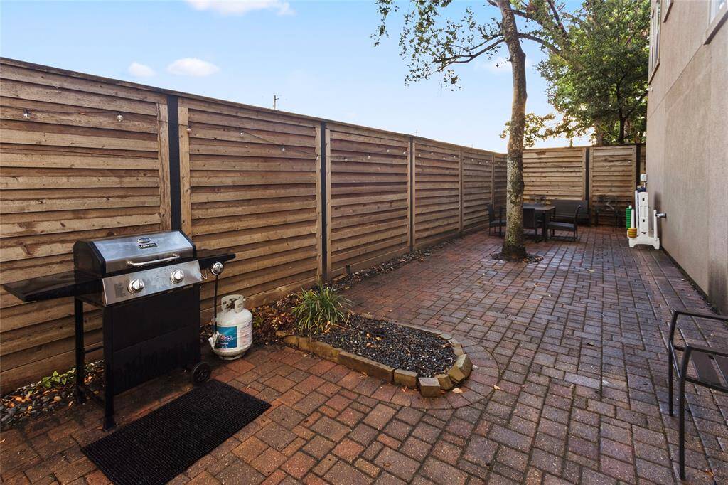 This photo showcases a private outdoor patio area with a brick-paved surface, featuring a wooden fence for privacy. The space includes a gas grill and outdoor seating, perfect for entertaining or relaxation. A tree adds a natural touch to the cozy atmosphere.