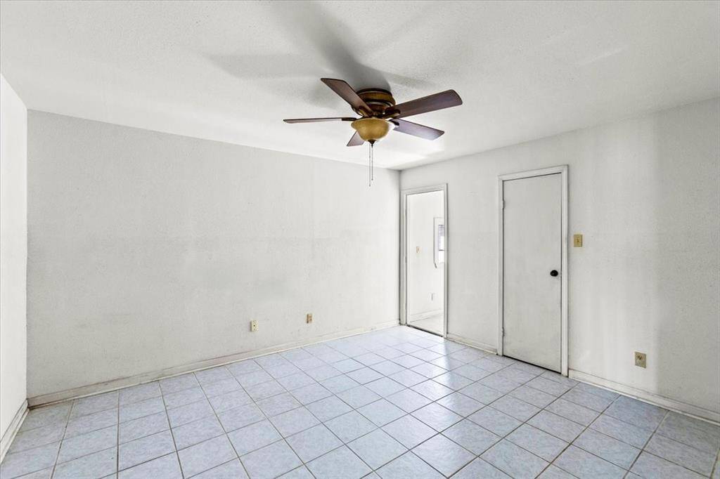 This primary bedroom is bright, featuring a ceiling fan and light tile flooring. There are two doors on the far wall, leading to an office space and a closet. The space offers a clean canvas for personalization.