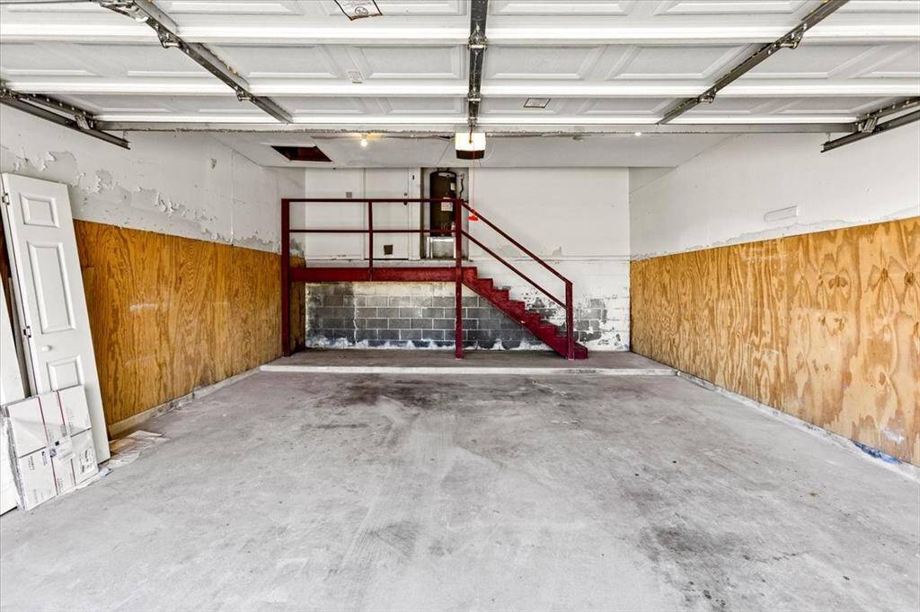 The photo shows a spacious, empty garage with a concrete floor, white walls partially covered with plywood, and a red staircase leading to the unit level. The garage door is automatic.