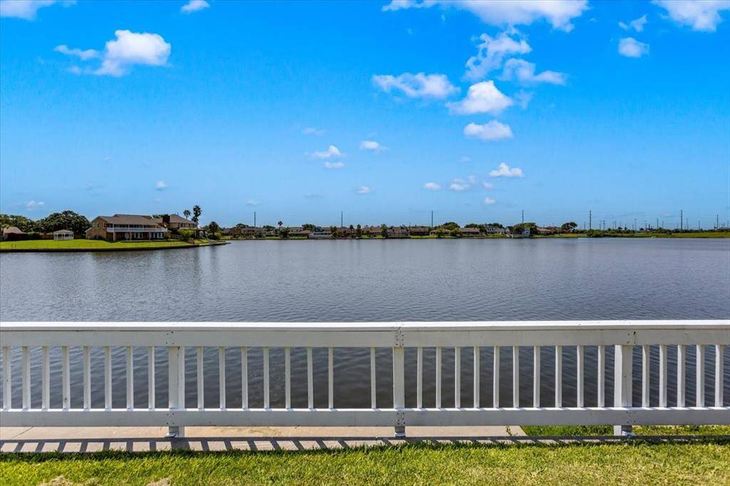 This photo showcases a serene waterfront view with a fence in the foreground, overlooking a calm lake with residential homes in the distance and a clear blue sky above.