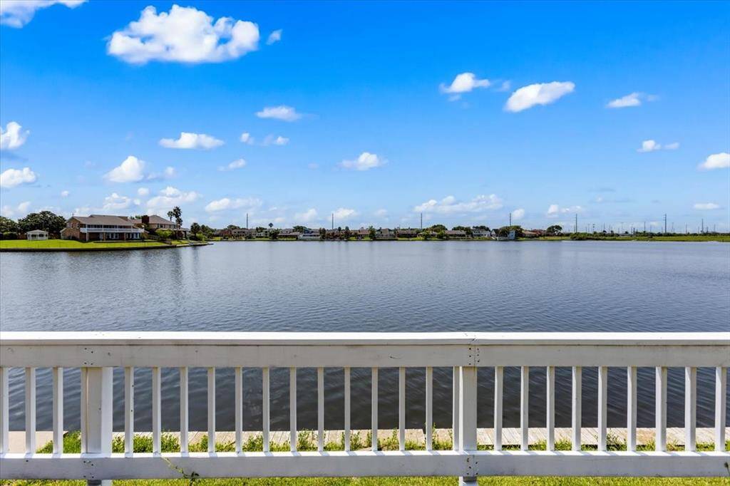 This photo showcases a serene waterfront view from a property with a white fence in the foreground, overlooking a calm lake with other houses in the distance under a blue sky dotted with fluffy clouds.