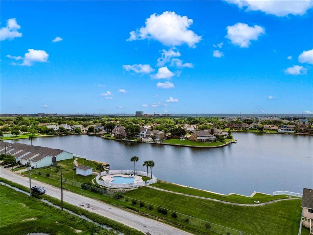 The photo shows a serene lakeside community with residential homes. Features include a communal swimming pool adjacent to the lake, well-manicured lawns, and a clear sky that suggests a peaceful living environment with attractive outdoor amenities.