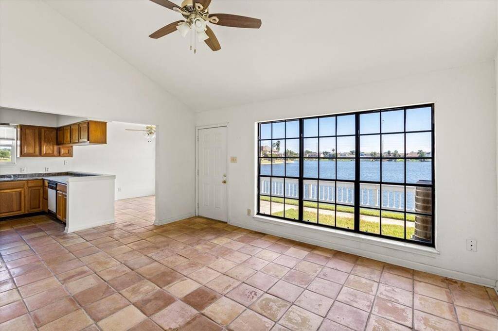 This is a bright, open-plan living space with a kitchen, featuring a large window with a scenic waterfront view, tile flooring, and a vaulted ceiling.