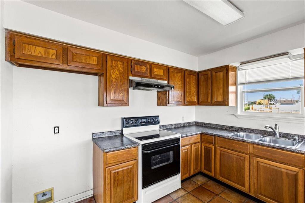 This kitchen features traditional wooden cabinetry, a built-in range hood, and a window overlooking the outside area. It has tile flooring and is equipped with a standard electric stove and a dual-basin sink.