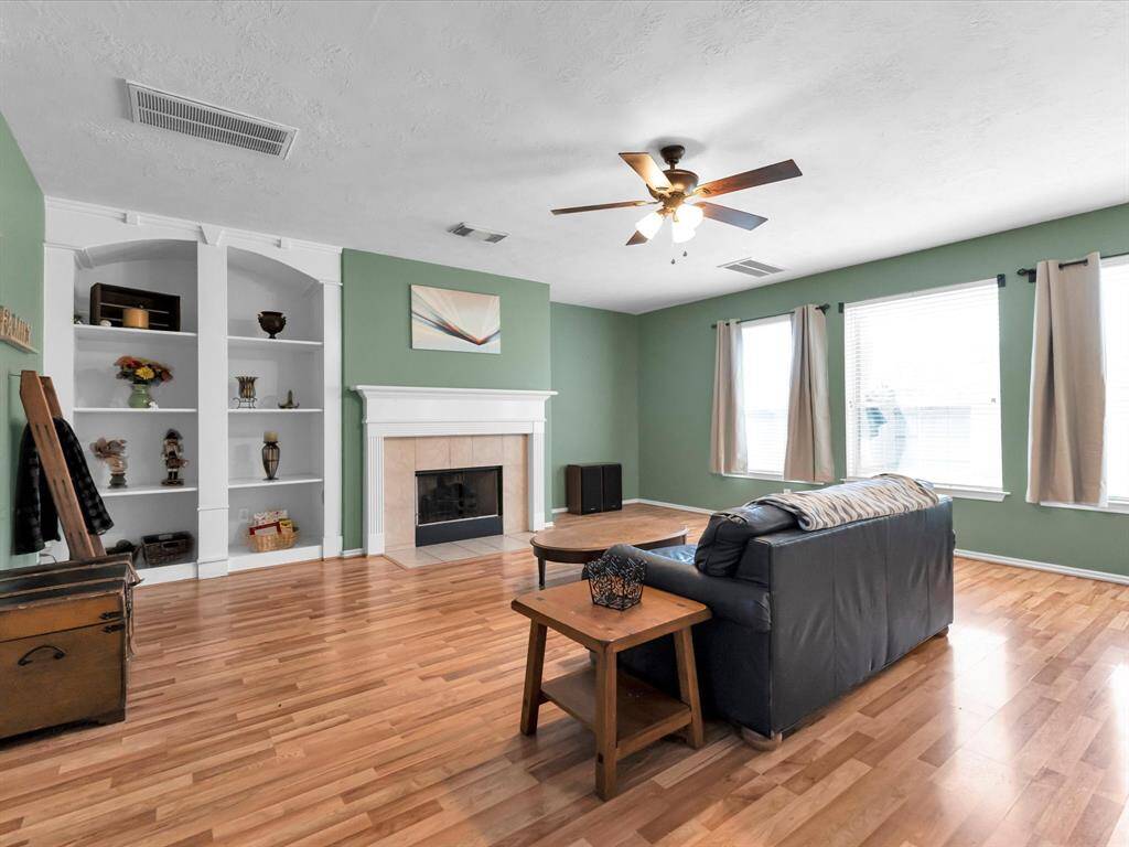 This is a spacious family room in a calming green hue with hardwood flooring, a central fireplace with a classic mantel, built-in shelving, and ample natural light from three large windows.