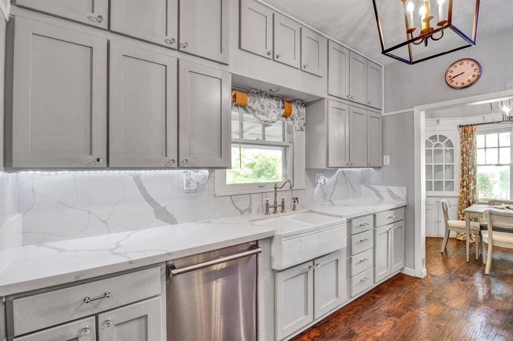 This is a well-lit, traditional kitchen featuring white cabinetry, stainless steel appliances, a farmhouse sink, and dark wood flooring. A unique chandelier adds character to the space.