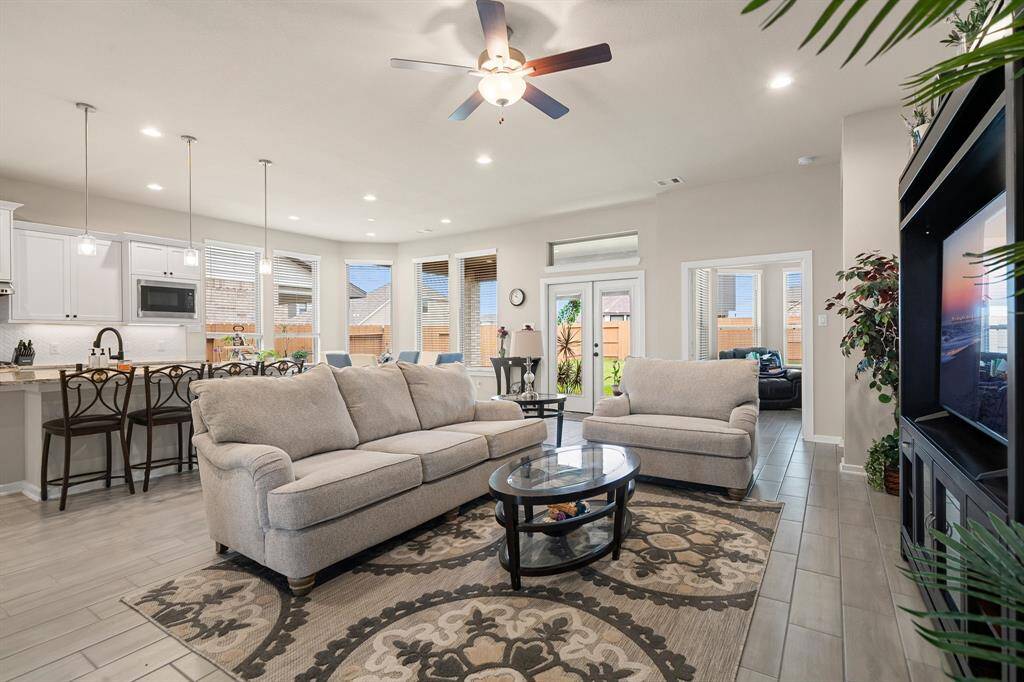 This is a spacious open-concept living space featuring a comfortable seating area, a modern kitchen with a breakfast bar, and ample natural light from the windows and doors leading to an outdoor space. It's well-lit with recessed lighting and a ceiling fan, and the interior has a contemporary feel with neutral tones and clean lines.