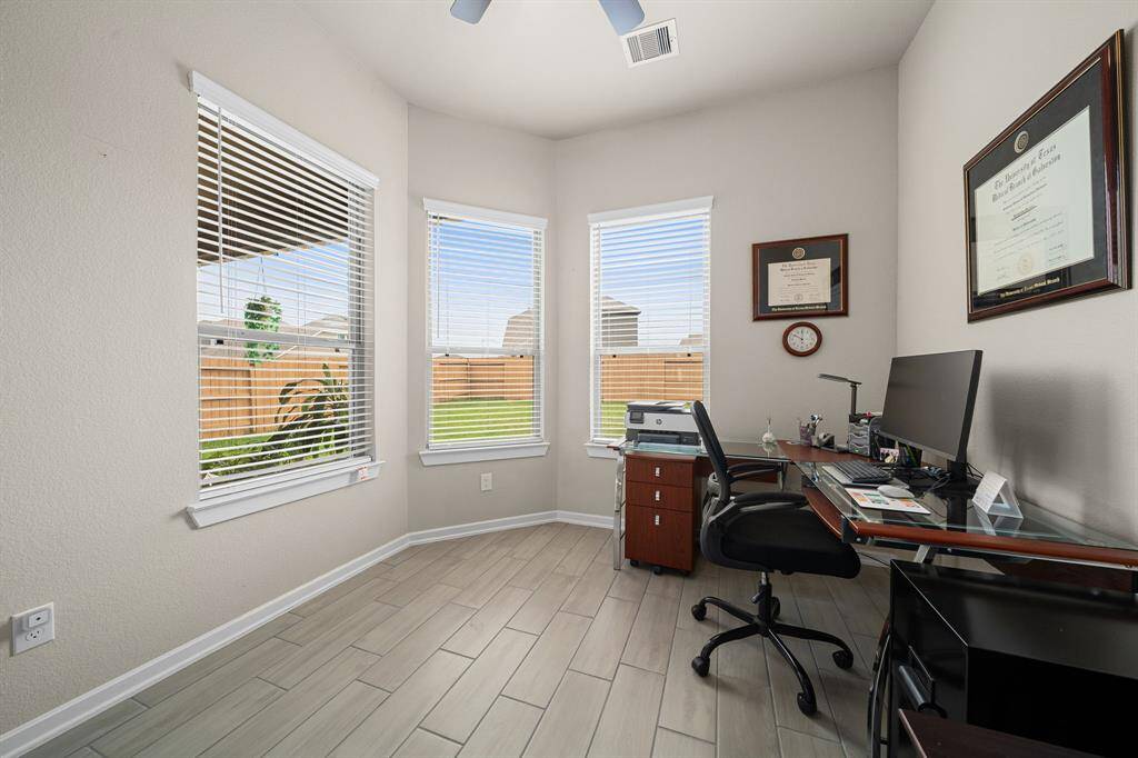 This is a bright, well-lit home office space featuring three large windows with blinds, modern flooring, and neutral wall colors.