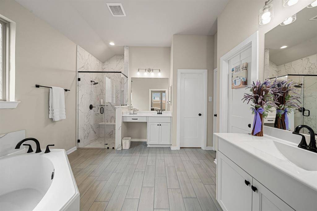 This is a spacious, modern bathroom featuring a double vanity with black fixtures, a large mirror, a walk-in shower with glass enclosure, and a separate bathtub. The room has light-colored wood-look flooring and marble-look tiles, creating a clean and elegant aesthetic.