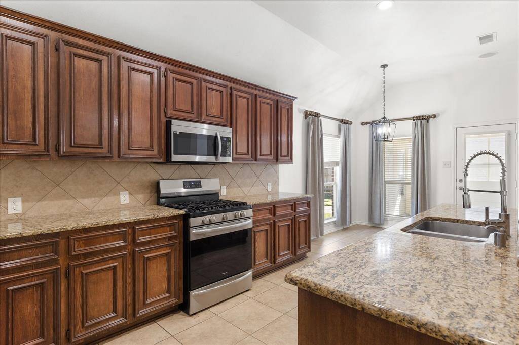 This is a spacious kitchen featuring rich, dark wood cabinetry, granite countertops, modern stainless steel appliances. It has a tiled backsplash and floor, with ample natural light coming through large windows. A stylish pendant light.