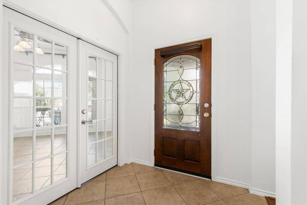 Mahogany front door with leaded-glass, on the right and white French doors on the left, leading to the study. The space is bright, with neutral-colored tile flooring and white walls, giving it a clean and open feel.