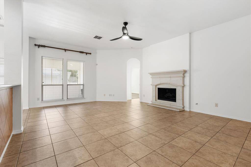 The photo shows a spacious and bright living room with tiled flooring, a fireplace, and a ceiling fan. Large windows allow for natural light, and the neutral color palette offers a blank canvas for personalization.