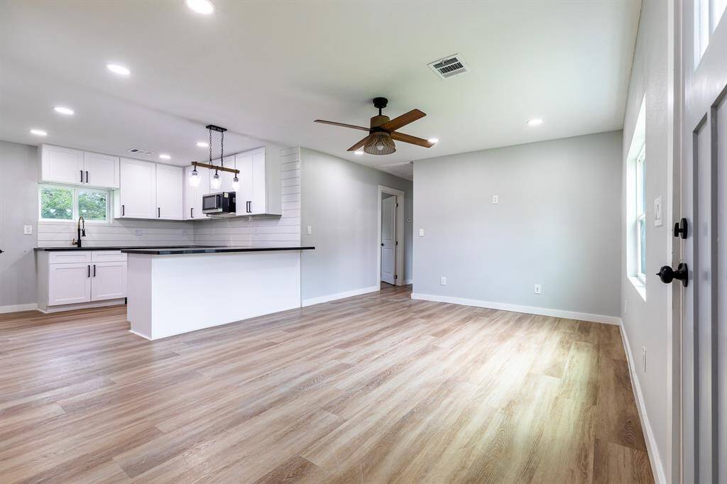 This is a bright, modern open-plan kitchen and living space featuring white cabinetry, stainless steel appliances, a large island with a sink, and wood flooring throughout. The area is well-lit with recessed lighting and a ceiling fan, and has a neutral color palette.