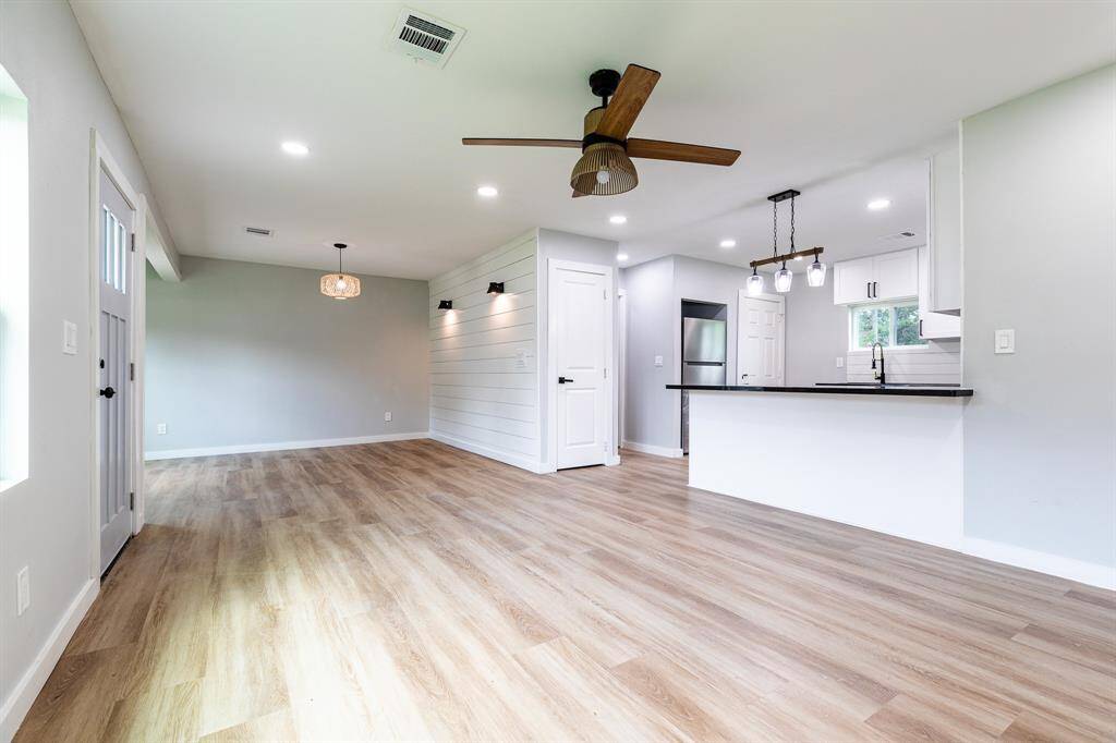 This is a modern, open-concept living space featuring wood flooring, fresh neutral paint, contemporary lighting fixtures, and a kitchen with white cabinetry and dark countertops. The room is well-lit with natural light and offers a welcoming atmosphere.