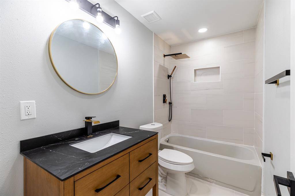 Modern bathroom featuring a wooden vanity with dark countertop, round mirror, stylish lighting, a combination bathtub and shower with sleek fixtures, and a white toilet.