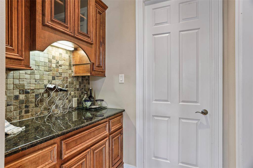 This kitchenette features rich wooden cabinetry, granite countertops, and a stylish tiled backsplash, complemented by under-cabinet lighting. There's also a white paneled door leading to another room.