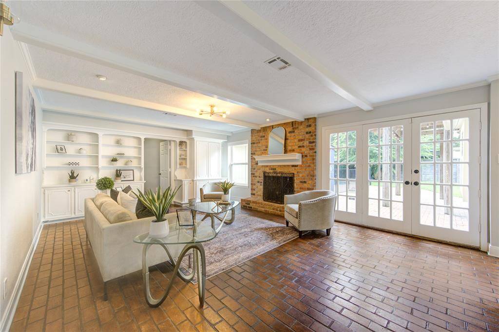 This spacious living room features a cozy brick fireplace, built-in shelving, and elegant French doors leading to the backyard. The room is bright and welcoming, with stylish furnishings and a mix of brick and carpet flooring. Perfect for relaxation and entertaining.