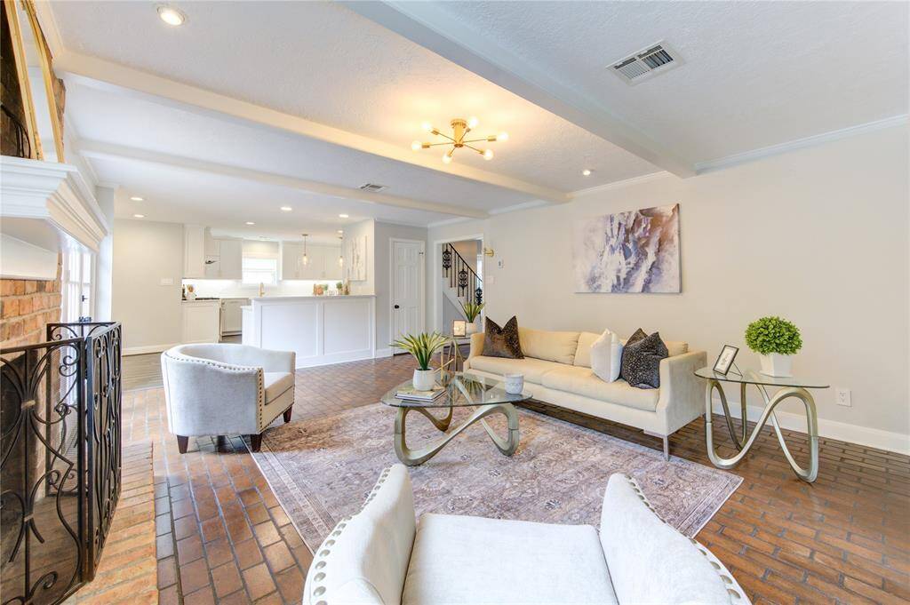 This inviting open-concept living area features a cozy fireplace, stylish furniture, and brick flooring. The bright, modern kitchen is seamlessly integrated, complete with white cabinets and a breakfast bar, creating a perfect space for entertaining.