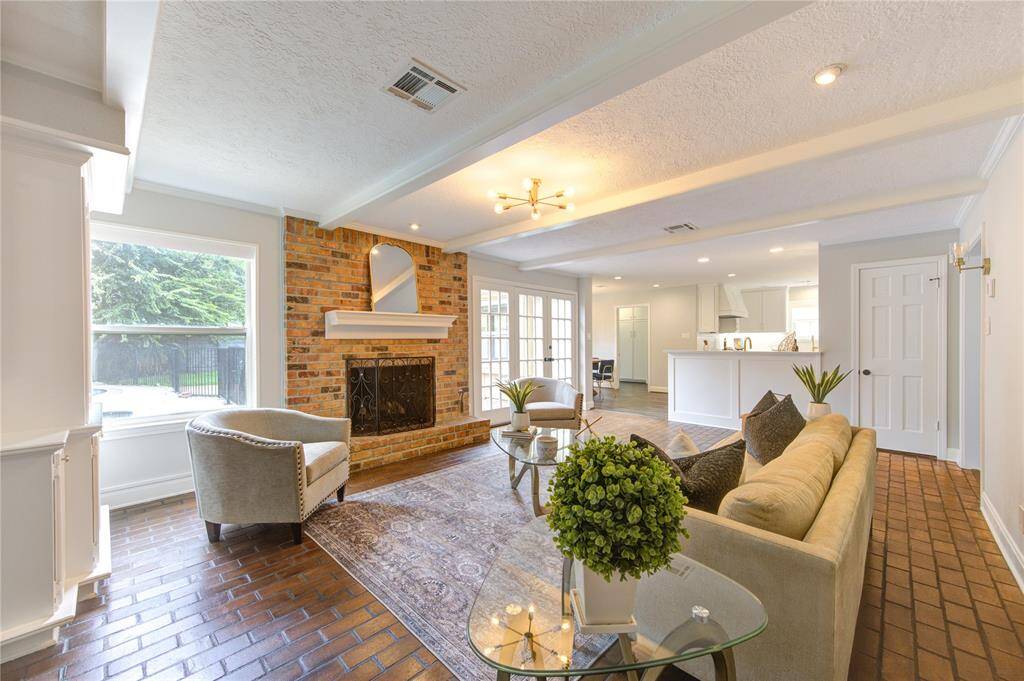 This inviting living room features a cozy brick fireplace, large windows for natural light, and elegant ceiling beams. The open layout flows into a modern kitchen, enhancing the spacious feel. Perfect for family gatherings and entertaining.