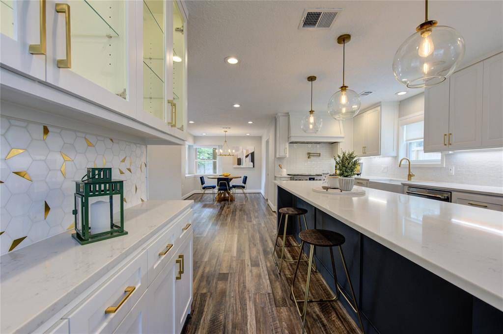 This modern kitchen features sleek white cabinetry, a long island with bar seating, and contemporary pendant lighting. The space is accented by a stylish hexagonal backsplash and rich wood flooring, offering a blend of elegance and functionality.