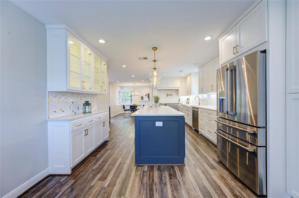 This modern kitchen features sleek stainless steel appliances, a spacious island with pendant lighting, and ample white cabinetry. The dark wood flooring adds warmth, while the open layout leads to a bright dining area.